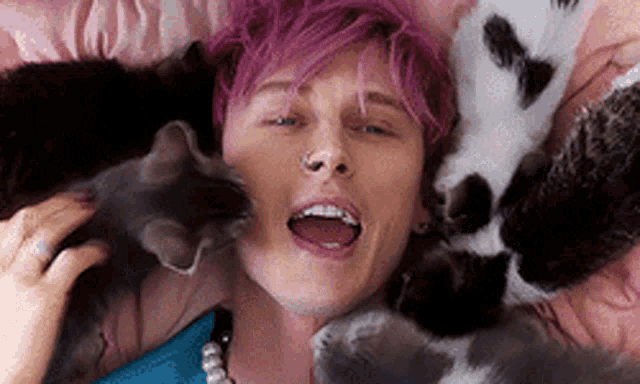 a man with pink hair is laying on a bed with three cats .