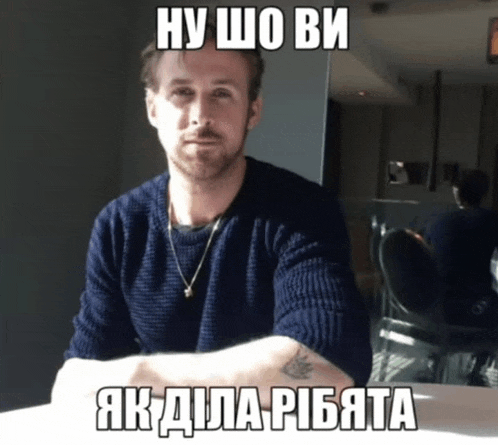 a man in a blue sweater sits at a table with his arms crossed in front of a sign that says ну шо ви
