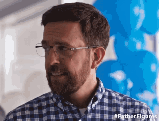 a man wearing glasses and a plaid shirt is standing in front of blue balloons and says father figures