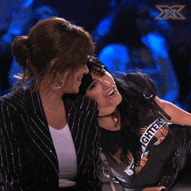 a woman kissing another woman on the cheek with a x-factor logo behind them