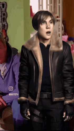 a man in a leather jacket is standing in a room with a woman in a purple jacket behind him .