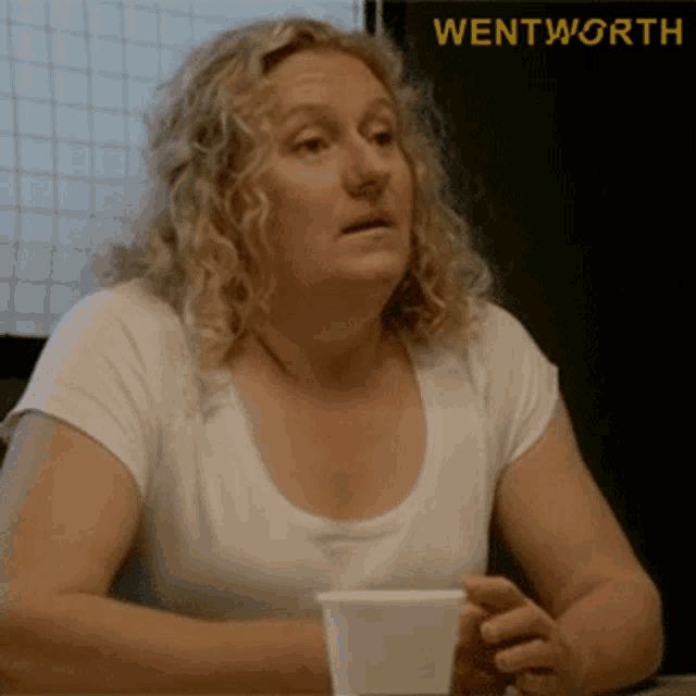 a woman in a white shirt is sitting at a table with a cup of coffee and the word wentworth on the bottom right