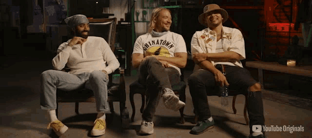 three men are sitting in chairs with one wearing a shirt that says downtown