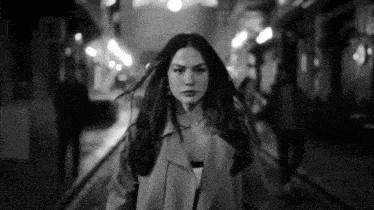 a black and white photo of a woman in a trench coat walking down a street at night .
