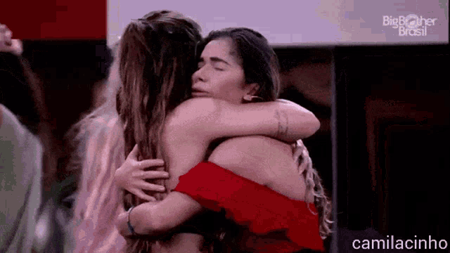 two women are hugging each other in a room . one is wearing a red top .
