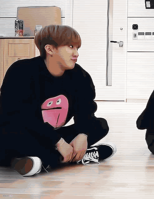 a young man wearing a black sweater with a pink face on it sits on the floor