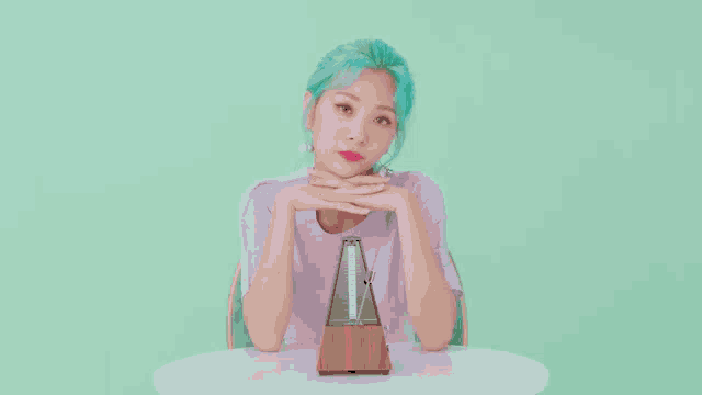 a woman with green hair is sitting at a table with her hands on her chin and a metronome in front of her .