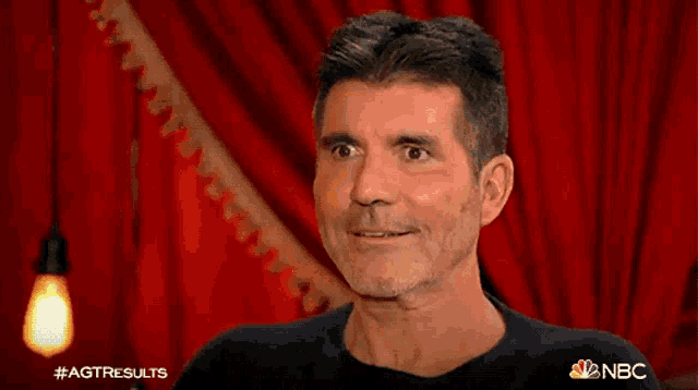 a man with a beard is sitting in front of a red curtain with the nbc logo on it .