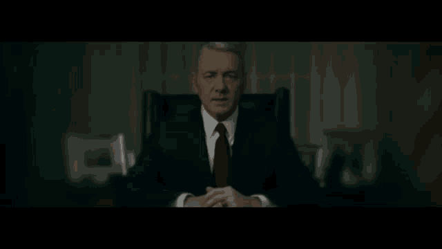 a man in a suit and tie is sitting in a dark room .