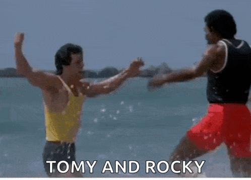 a man is hugging another man on the beach with the words `` tomy and rocky '' written below him .