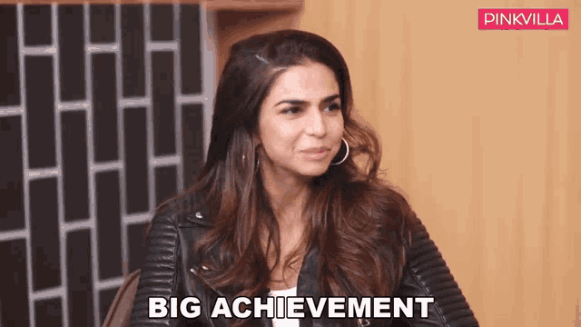 a woman in a leather jacket says big achievement in front of a brick wall