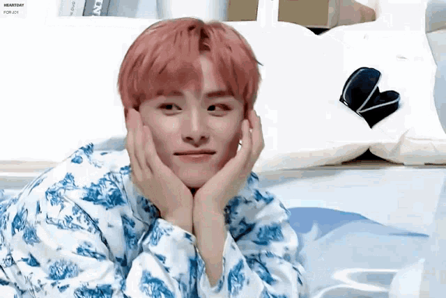 a man with pink hair is laying on a bed with his hands on his face .