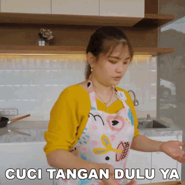 a woman wearing a pink apron is standing in a kitchen with the words cuci tangan dulu ya below her