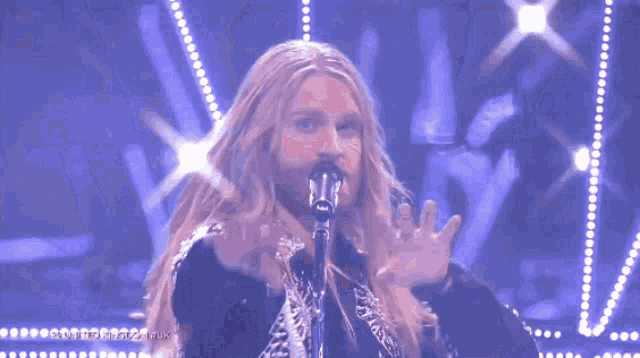 a man with long blonde hair is singing into a microphone with a purple background