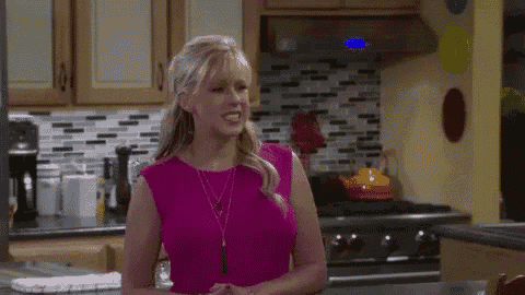 a woman in a pink top is standing in a kitchen with her hands folded .