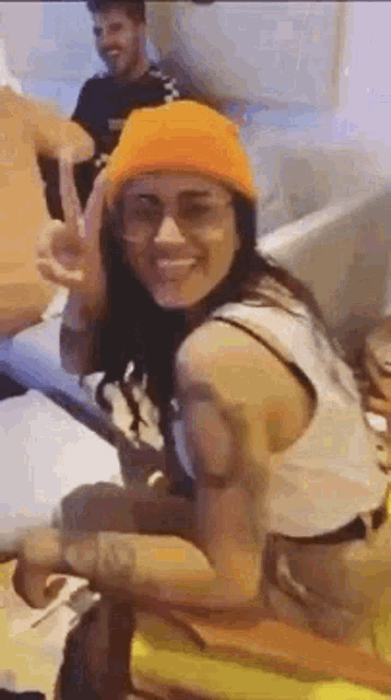 a woman wearing an orange beanie and glasses is sitting on a couch giving a peace sign .