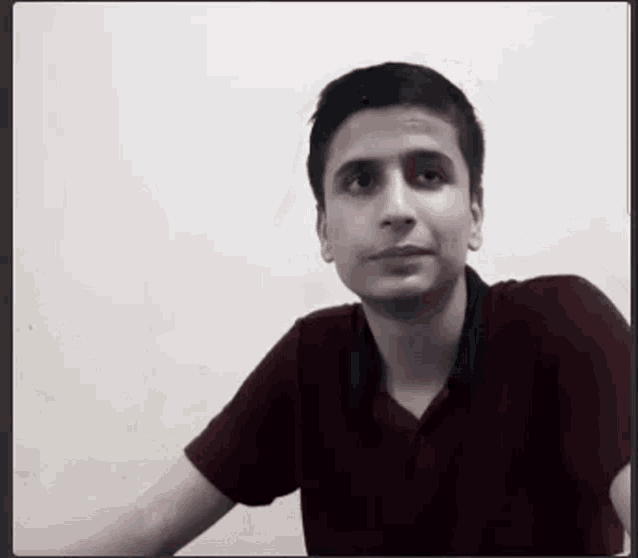a young man in a maroon shirt is sitting in front of a white wall and looking at the camera .