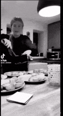 a black and white photo of a woman cooking in a kitchen with plates of food on the counter