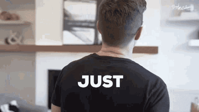 a man wearing a black t-shirt with the word just on the back is standing in front of a fireplace .