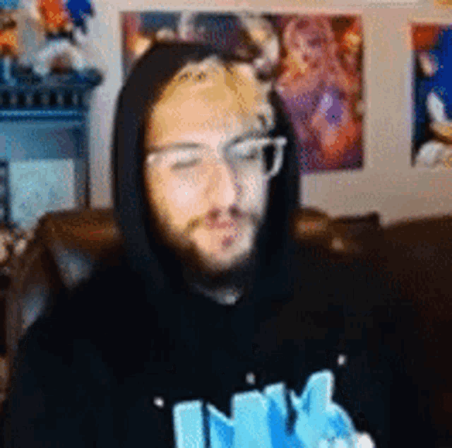 a man with a beard and glasses is wearing a black hoodie and a blue shirt .
