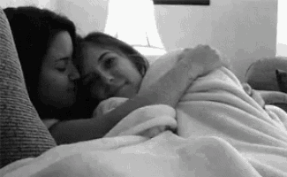 two women are hugging each other while laying on a couch under a blanket .