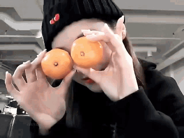 a woman in a black hat is holding two oranges in front of her face .