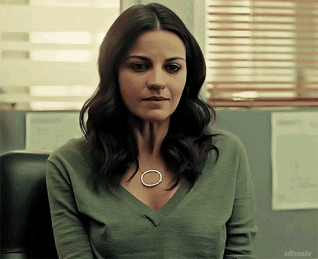 a woman wearing a green sweater is sitting in front of a window with blinds on it