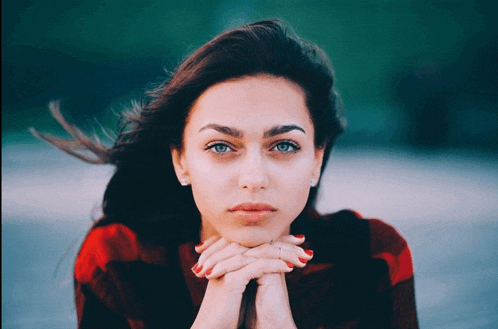 a woman with her hands folded looks at the camera with her hair blowing in the wind