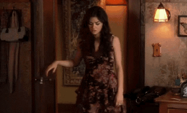 a woman in a floral dress is standing in a room with a lamp on the wall