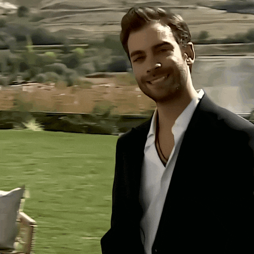 a man in a suit and white shirt smiles in front of a grassy field