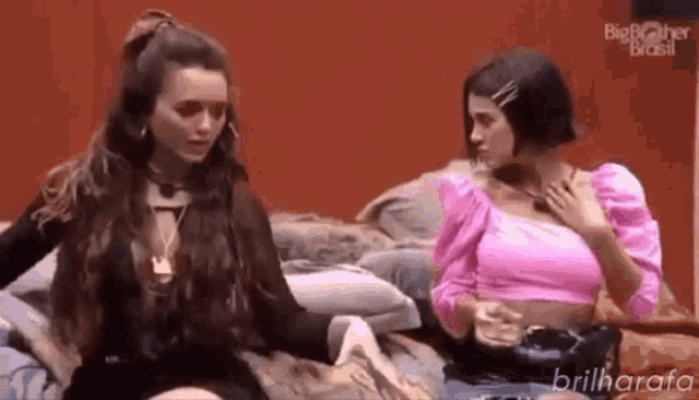 two women are sitting on a bed talking to each other . one of the women is wearing a pink top .