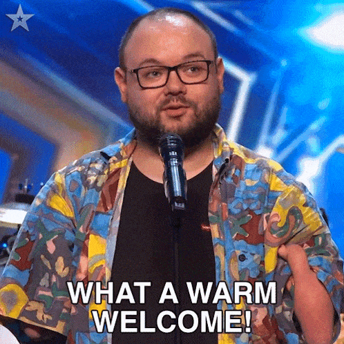 a man wearing glasses is standing in front of a microphone and saying what a warm welcome !