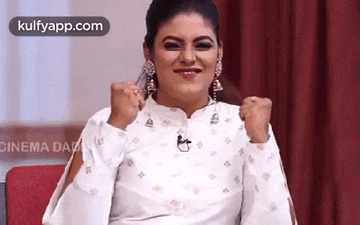 a woman in a white shirt and earrings is sitting on a red couch with her hands in the air .