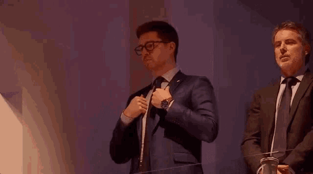 a man in a suit and tie is adjusting his tie while standing next to another man in a suit and tie .