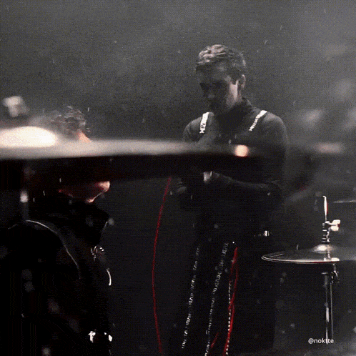 a man in a black turtleneck stands in front of a drum set with the hashtag @noktee on the bottom
