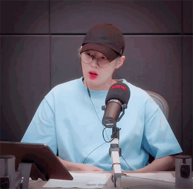 a man wearing a hat and glasses is sitting in front of a microphone with the word now on it
