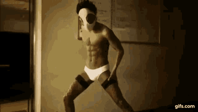 a shirtless man wearing a mask and white underwear is dancing in a dark room .
