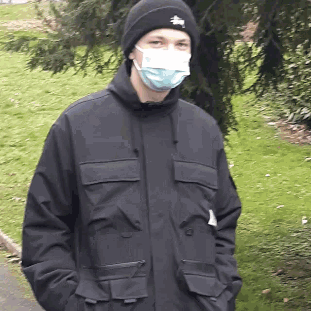 a man wearing a mask and a black jacket is standing in a park