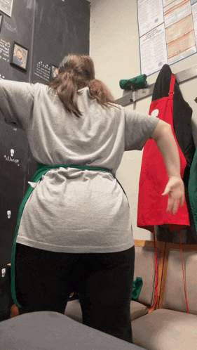 a woman in a gray shirt and green apron is standing in front of a wall with a sign that says " welcome " on it