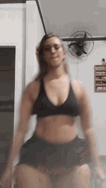 a woman in a black bra and shorts is dancing in a room with a fan on the wall .