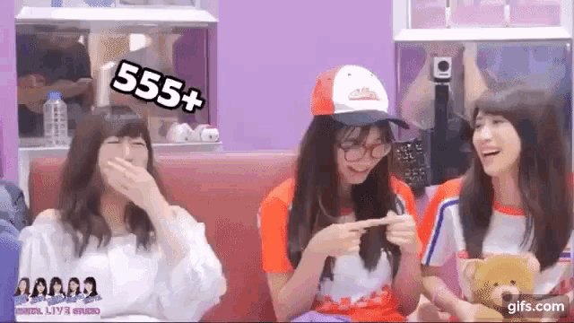 three girls are sitting on a couch and one of them is wearing a hat with the number 555+ on it .