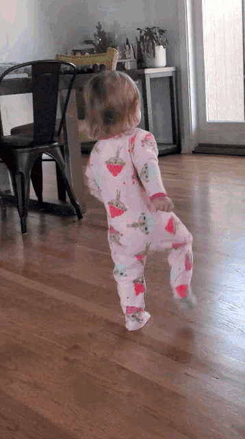 a little girl in a pink pajamas is walking on a wood floor