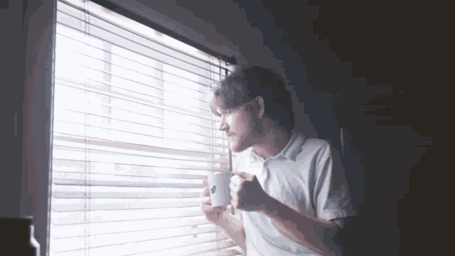 a man looking out a window while holding a coffee mug with a blue heart on it