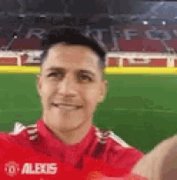 a man in a red shirt is taking a selfie with his hand in front of a soccer field .