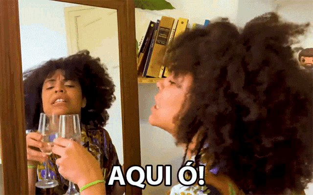 a woman with curly hair holds a glass of wine in front of a mirror and says aqui o!