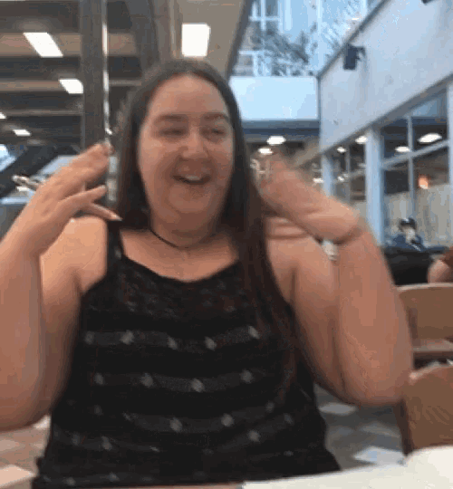 a woman in a black tank top is sitting at a table with her hands up