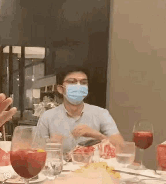 a man wearing a face mask is sitting at a table with glasses of wine and food .