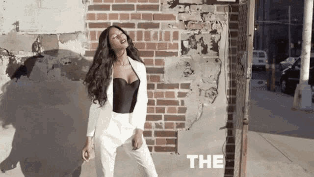 a woman is standing in front of a brick wall wearing a white suit and a black top .