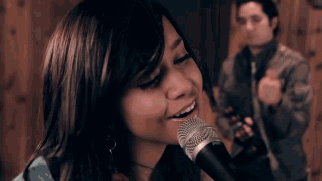 a woman singing into a shure microphone with a man playing a guitar in the background