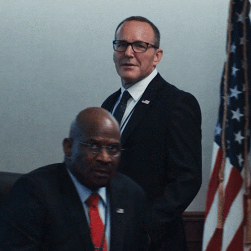 a man in a suit and tie stands next to another man with glasses